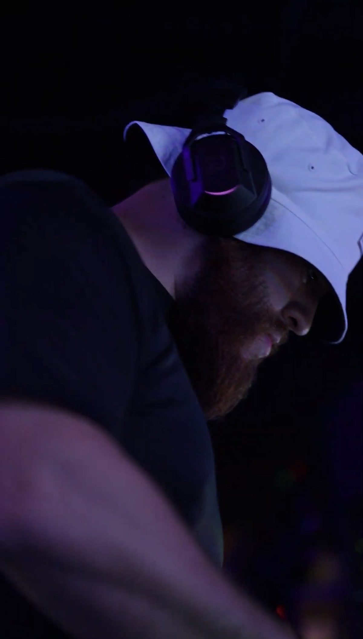 G in a white bucket hat and headphones, in profile under purple light